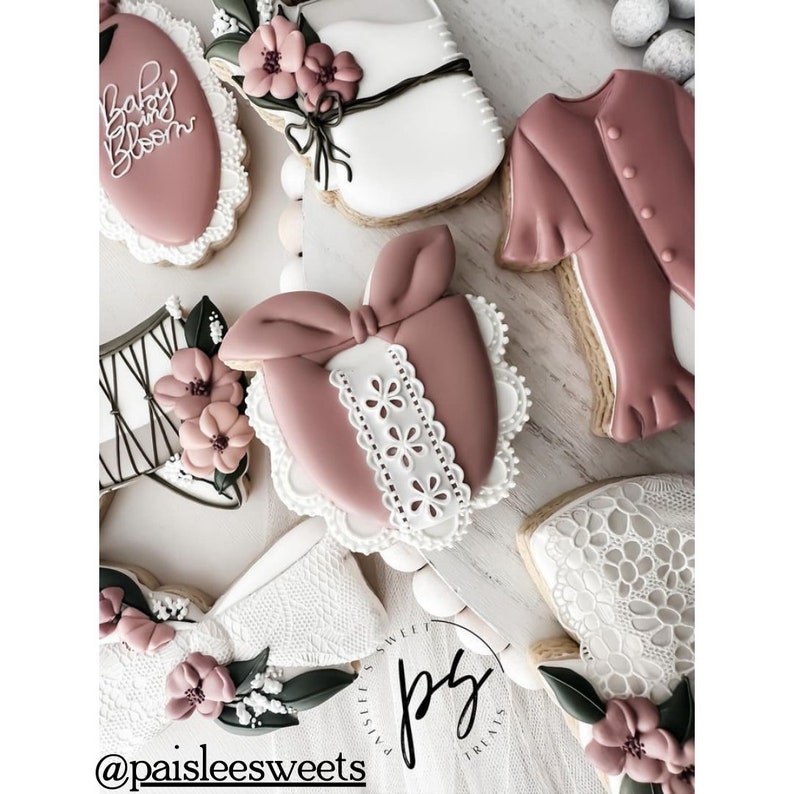 May include: A collection of decorated sugar cookies with various designs, including a baby onesie, a bow, a flower, and a lace pattern. The cookies are decorated in shades of pink, white, and green. The cookies are arranged on a white surface. The text "Baby Bloom" is visible on one of the cookies.