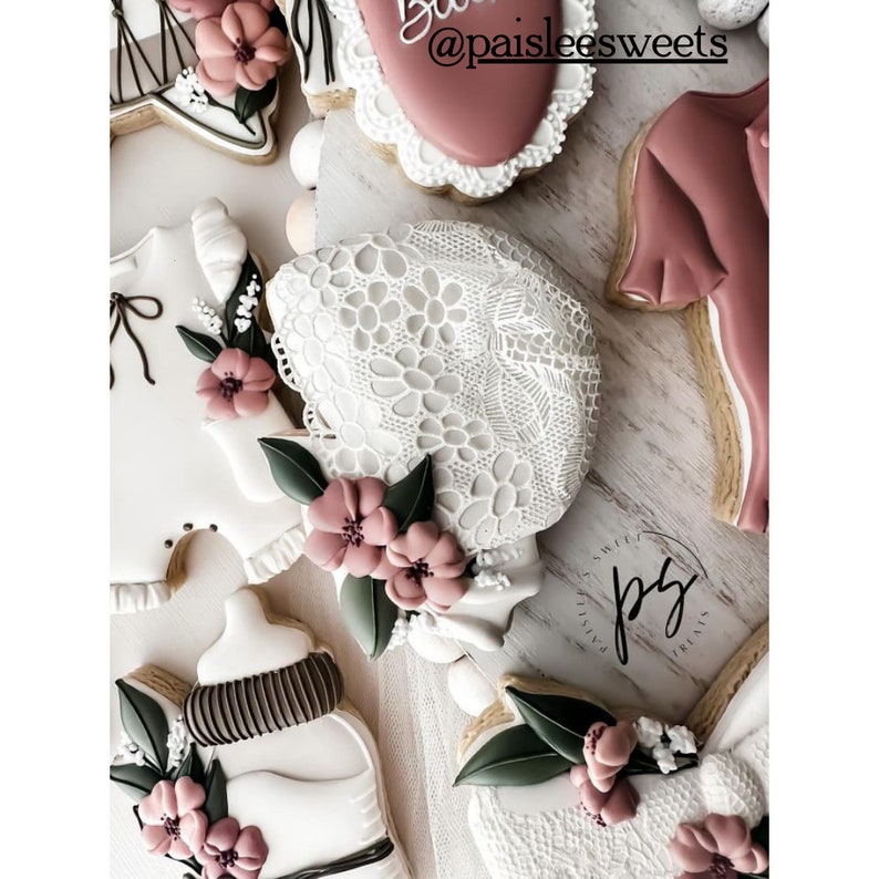 May include: A set of decorated sugar cookies with a baby theme. The cookies are decorated with white lace, pink flowers, and green leaves. One cookie is shaped like a baby bottle, another is shaped like a baby onesie, and another is shaped like a bonnet. The cookies are decorated with the text "@paisleesweets".