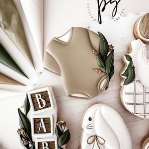 K&ouml;nnte beinhalten: Ein Set dekorierter Zuckerkekse mit Baby-Motiv. Die Kekse sind mit wei&szlig;em Zuckerguss verziert und zeigen einen Baby-Strampler, ein Kaninchen, einen Stapel Bl&ouml;cke, die "BABY" buchstabieren, und ein Karomuster. Die Kekse sind von Spritzbeuteln und einem Logo mit der Aufschrift "Paislee's Sweet Treats" umgeben.