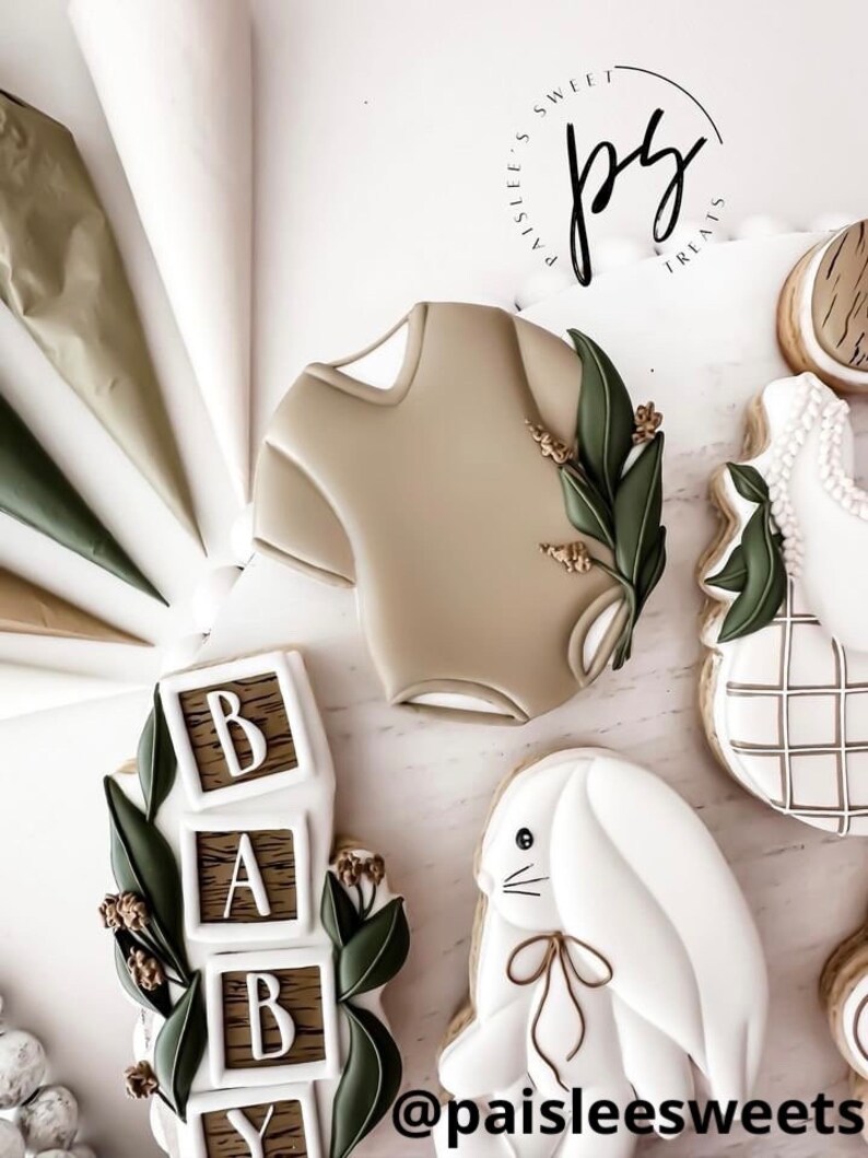 K&ouml;nnte beinhalten: Ein Set dekorierter Zuckerkekse mit Baby-Motiv. Die Kekse sind mit wei&szlig;em Zuckerguss verziert und zeigen einen Baby-Strampler, ein Kaninchen, einen Stapel Bl&ouml;cke, die "BABY" buchstabieren, und ein Karomuster. Die Kekse sind von Spritzbeuteln und einem Logo mit der Aufschrift "Paislee's Sweet Treats" umgeben.