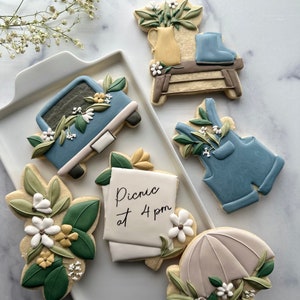 May include: A set of six decorated sugar cookies featuring a blue truck, a pair of blue overalls, a floral arrangement in a yellow pitcher, a wooden stool, a note that says "Picnic at 4 pm", and a white umbrella with green leaves and white flowers.