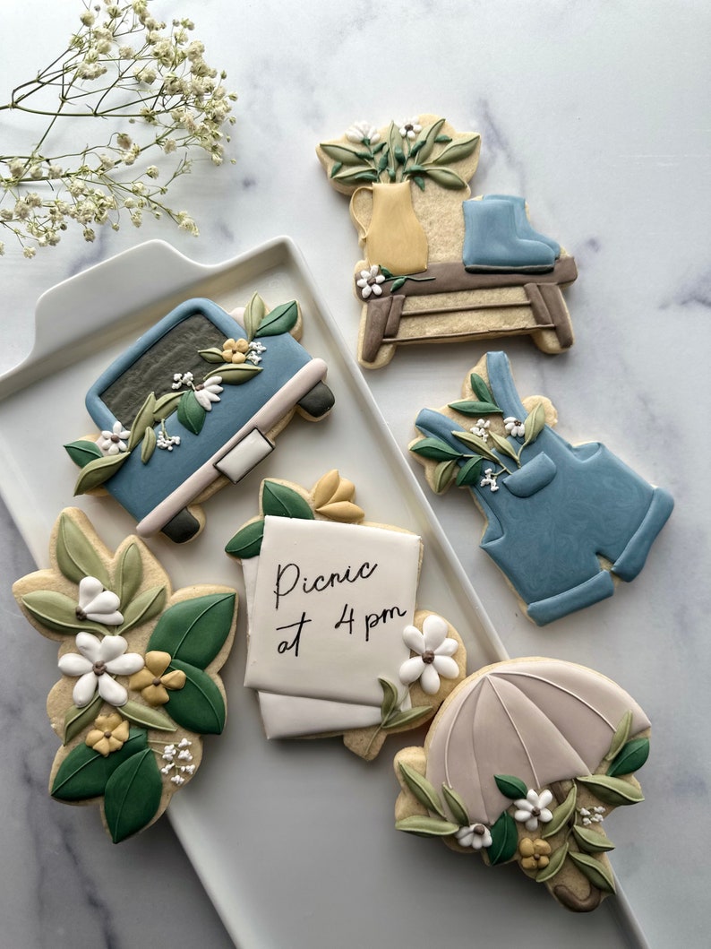May include: A set of six decorated sugar cookies featuring a blue truck, a pair of blue overalls, a floral arrangement in a yellow pitcher, a wooden stool, a note that says "Picnic at 4 pm", and a white umbrella with green leaves and white flowers.