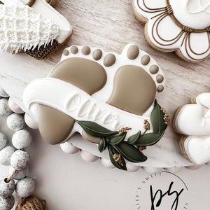 K&ouml;nnte beinhalten: Ein verzierter Zuckerkeks mit einem Paar Babyf&uuml;&szlig;en, einem Banner mit dem Namen "Oliver" und gr&uuml;nen Bl&auml;ttern. Der Keks befindet sich auf einem wei&szlig;en Hintergrund mit einem braunen und wei&szlig;en Quast und einer wei&szlig;en und braunen Perlenkette.