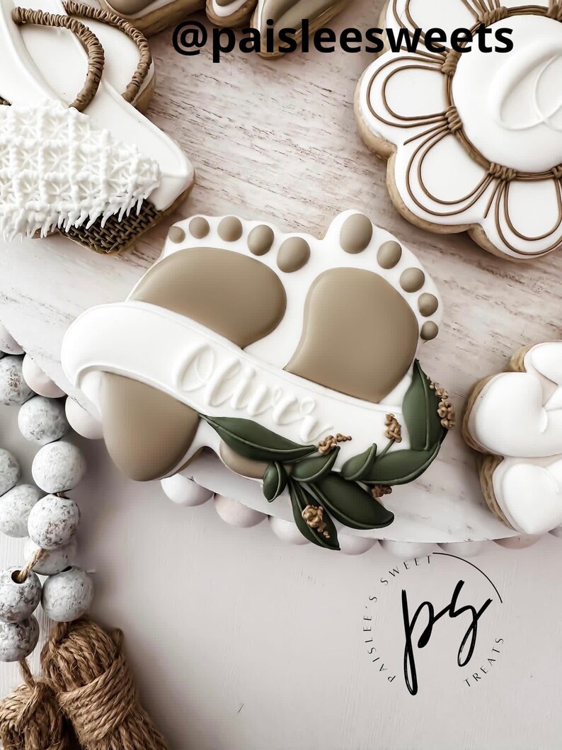 K&ouml;nnte beinhalten: Ein verzierter Zuckerkeks mit einem Paar Babyf&uuml;&szlig;en, einem Banner mit dem Namen "Oliver" und gr&uuml;nen Bl&auml;ttern. Der Keks befindet sich auf einem wei&szlig;en Hintergrund mit einem braunen und wei&szlig;en Quast und einer wei&szlig;en und braunen Perlenkette.