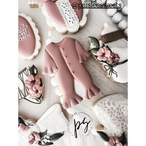 May include: A set of six decorated sugar cookies featuring a baby onesie, a baby bottle, a floral design, and a lace design. The cookies are decorated in shades of pink, white, and green.