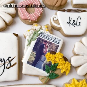 May include: A decorated sugar cookie with a printed image of a wedding couple and the text "BREAKING NEWS THE KELLEY'S FIRST ANNIVERSARY". The cookie is decorated with white daisies, yellow sunflowers, and green leaves.