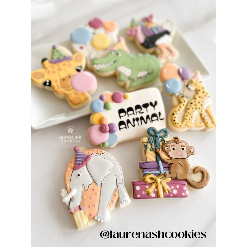 K&ouml;nnte beinhalten: Ein Set dekorierter Zuckerkekse mit Tiermotiven, darunter eine Giraffe, ein Krokodil, ein Gepard, ein Elefant und ein Affe. Die Kekse sind mit buntem Zuckerguss und Streuseln verziert. Ein Keks tr&auml;gt den Text "Party Animal".