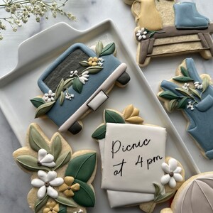 May include: A set of six decorated sugar cookies featuring a blue truck with flowers, a floral arrangement in a yellow pitcher, a pair of blue boots, a note that says "Picnic at 4 pm", a floral arrangement, and a light blue umbrella.