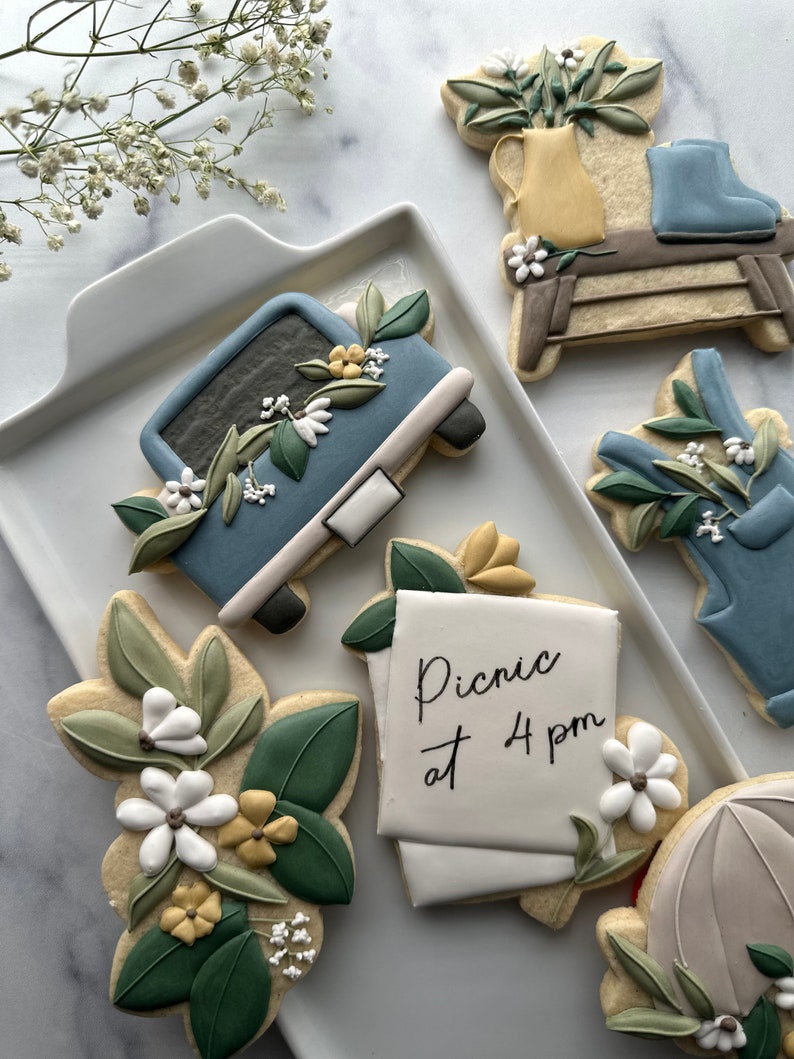 May include: A set of six decorated sugar cookies featuring a blue truck with flowers, a floral arrangement in a yellow pitcher, a pair of blue boots, a note that says "Picnic at 4 pm", a floral arrangement, and a light blue umbrella.