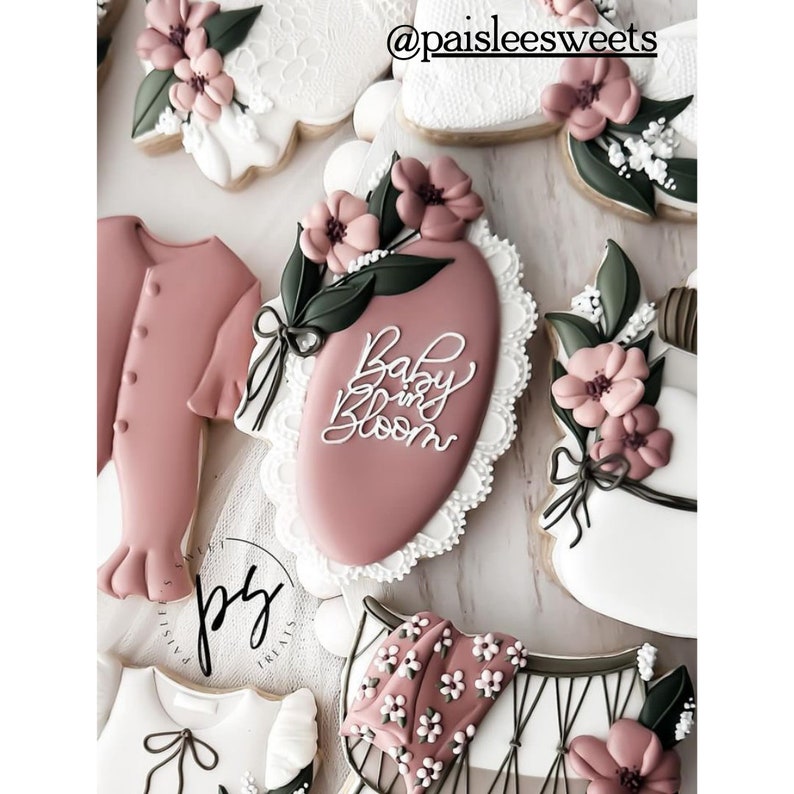 May include: A pink and white decorated sugar cookie with the text "Baby in Bloom" in white icing. The cookie is decorated with a floral design and lace trim.