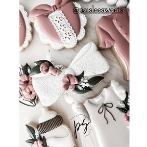 May include: A set of decorated sugar cookies featuring a pink bow, a pink heart with lace trim, a pink shirt, a white baby bottle, and a white onesie with a brown bow. The cookies are decorated with white lace, pink flowers, and green leaves.