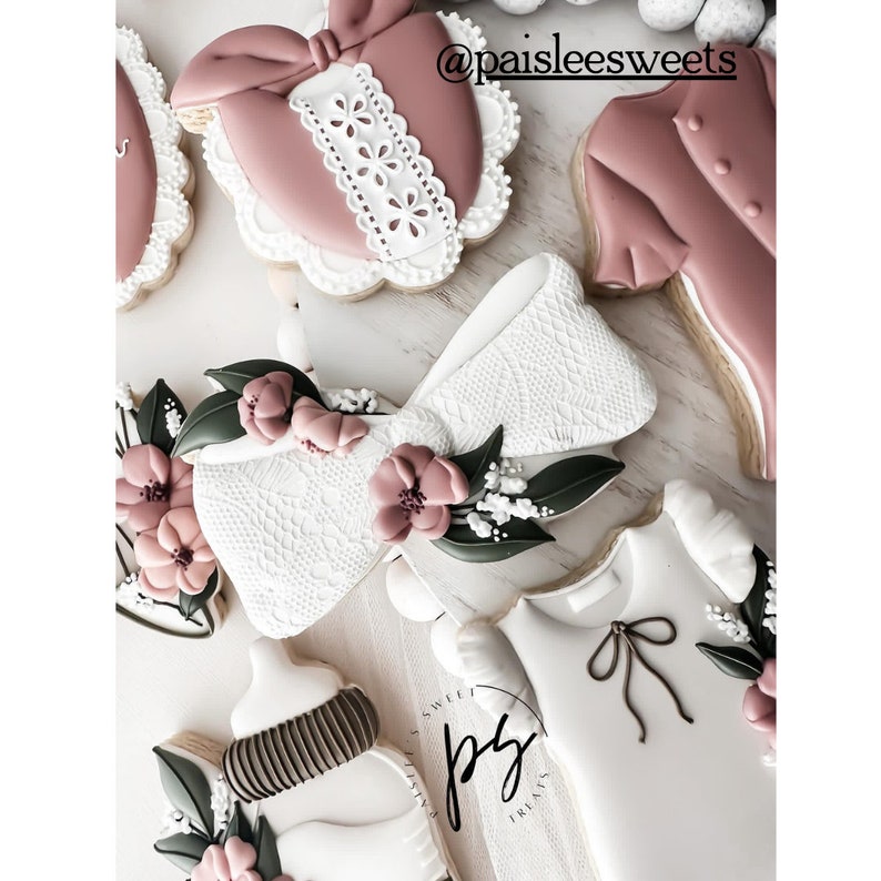 May include: A set of decorated sugar cookies featuring a pink bow, a pink heart with lace trim, a pink shirt, a white baby bottle, and a white onesie with a brown bow. The cookies are decorated with white lace, pink flowers, and green leaves.