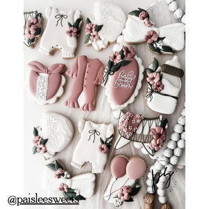May include: A collection of decorated sugar cookies featuring baby-themed designs, including onesies, a crib, a bottle, and a banner that reads "Baby Bloom". The cookies are decorated with pink, white, and green icing and have a delicate, feminine aesthetic.