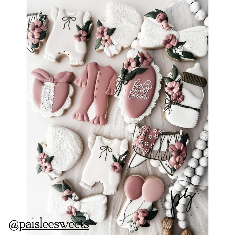 May include: A collection of decorated sugar cookies featuring baby-themed designs, including onesies, a crib, a bottle, and a banner that reads "Baby Bloom". The cookies are decorated with pink, white, and green icing and have a delicate, feminine aesthetic.