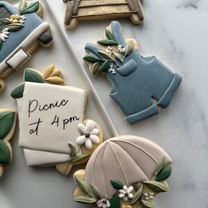 May include: A set of five decorated sugar cookies. The cookies are decorated with blue, white, and green icing. One cookie has the text "Picnic at 4 pm" written on it. Another cookie is shaped like an umbrella with a floral design.