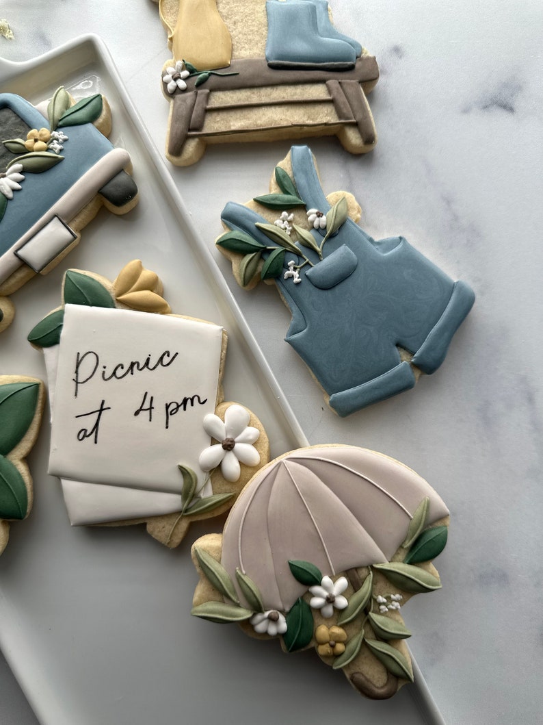 May include: A set of five decorated sugar cookies. The cookies are decorated with blue, white, and green icing. One cookie has the text "Picnic at 4 pm" written on it. Another cookie is shaped like an umbrella with a floral design.