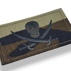 May include: A black skull and crossbones patch with a camouflage background. The patch is made of fabric and has a stitched edge.