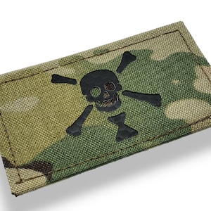 Puede incluir: Un parche de calavera y huesos cruzados en blanco y negro sobre un fondo de tela de camuflaje.