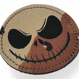 Puede incluir: Un parche redondo de tela de camuflaje marrón y beige con una silueta negra de una calavera sonriente con la boca y los ojos cosidos.