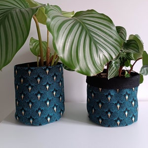 Puede incluir: Dos macetas decorativas con plantas verdes y frondosas. Las macetas son cilíndricas y presentan un estampado turquesa y negro con detalles dorados. Las plantas tienen hojas grandes y verdes a rayas. Las macetas están sobre una superficie blanca.