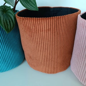 Op de afbeelding: Drie cilindrische plantenpotten van stof in turquoise, oranje en roze. De potten hebben een corduroy textuur en zijn gevoerd met zwarte stof.
