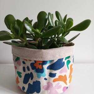 Op de afbeelding: Een witte stoffen plantenbak met een kleurrijk abstract bloemenpatroon. De plantenbak is gevuld met een groene vetplant.
