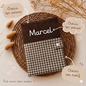 Peut inclure: Une pochette rectangulaire composée d'une partie supérieure en velours côtelé marron foncé avec le prénom Marcel brodé en blanc, et d'une partie inférieure à motif vichy marron et blanc. Un petit bouton-pression blanc est visible sur le devant.