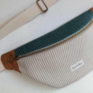 Op de afbeelding: Een beige, bruin en turquoise heuptas met een ritssluiting en een bruin leren band. De heuptas heeft een wit label met de tekst "Sarenko" erop.