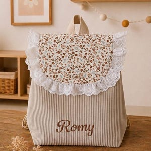 Peut inclure: Un sac à dos en velours côtelé beige avec un rabat à motif floral marron et vert, bordé de dentelle anglaise blanche. Le prénom Romy est brodé en lettres cursives marron sur le devant du sac, qui possède une poignée et des bretelles.
