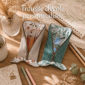 May include: Two corduroy pencil cases with zipper closures and fabric bow accents. One case is beige with a floral interior lining containing pens, and the other is teal with a green botanical interior lining holding a pair of eyeglasses.