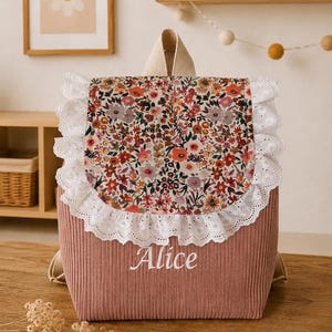 Peut inclure: Un sac à dos en velours côtelé rose poudré avec un rabat à motif floral bordé de dentelle blanche à œillets. Le prénom Alice est brodé en blanc sur la poche avant. Le sac possède des bretelles beiges et une poignée supérieure de transport.