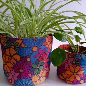 Könnte beinhalten: Zwei Topfpflanzen in bunten, floralen Stoffgefäßen. Der größere Topf ist blau mit orangefarbenen, rosa und gelben Blumen. Der kleinere Topf ist orange mit rosa, blauen und gelben Blumen.