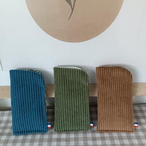Peut inclure: Trois étuis en velours côtelé, bleu sarcelle, vert olive et marron, chacun avec un petit détail de drapeau français. Les étuis sont rectangulaires avec des dessus arrondis, et sont exposés sur une surface à carreaux. Les étuis sont probablement pour des lunettes ou des téléphones.
