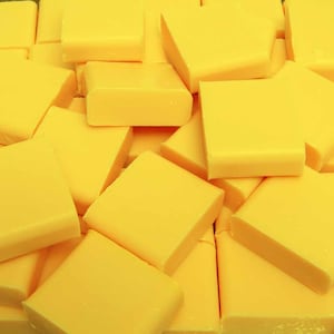 May include: A close-up shot of a pile of bright yellow cheese blocks. The cheese blocks are rectangular with slightly rounded edges, and they are stacked together. The lighting is bright, and the cheese appears fresh.