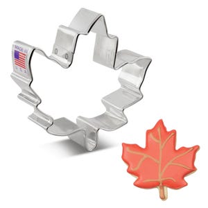 May include: A silver metal maple leaf cookie cutter with a red and orange maple leaf cookie. The cookie cutter is labeled "Made in U.S.A."