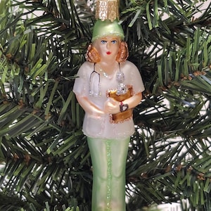 May include: A glass Christmas ornament depicting a nurse wearing a green scrub top and pants. She is holding a clipboard and a stethoscope. The ornament has a gold top and is decorated with glitter.