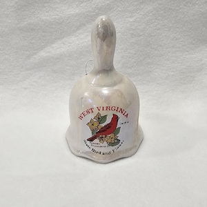 May include: A pearlescent ceramic bell with a handle and a small loop for hanging. The bell features a graphic of a red cardinal bird and the text "West Virginia State Bird and Flower".