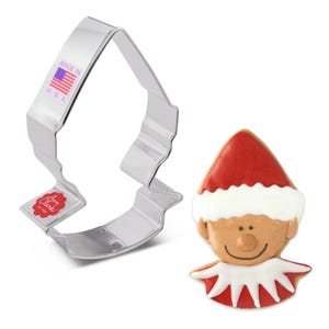 May include: A metal cookie cutter shaped like an elf's head, with a red hat, white trim, and a smiling face. The cutter is made in the USA and is next to a decorated gingerbread cookie shaped like an elf.