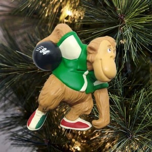 May include: A brown monkey wearing a green and white bowling shirt and holding a black bowling ball with the number "94" on it. The monkey is wearing red and white bowling shoes. The monkey is a Christmas ornament.