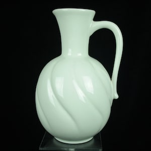Op de afbeelding: Een witte keramische kan met een draaiend ontwerp en een handvat. De kan heeft een rond lichaam en een smalle hals.