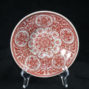 Può includere: Un piatto decorativo in ceramica con un motivo floreale rosso e bianco. Il piatto presenta un fiore centrale circondato da altri motivi floreali. È esposto su un supporto in acrilico trasparente su sfondo nero.