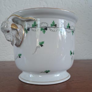 Op de afbeelding: Een witte porseleinen plantenpot met een gebeeldhouwd geitenkop-handvat met gouden accenten. De buitenkant is versierd met een herhalend patroon van groene en zwarte klaverachtige bladeren en dunne, krullende rankmotieven rond de bovenrand.