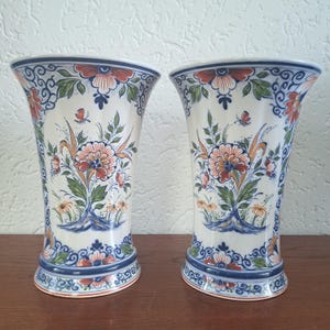 May include: A pair of matching ceramic vases with a flared rim and base. The white surface features hand-painted floral motifs in shades of orange, green, and blue, with intricate scrollwork patterns decorating the top and bottom borders.