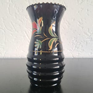 May include: Black glass vase with a flared top and a ribbed base. The vase features hand-painted floral designs in red, green, and gold. The rim has a scalloped edge. A decorative piece for home decor.