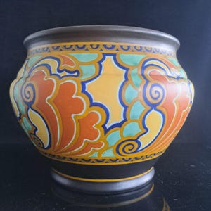 Può includere: Una ciotola in ceramica con base e bordo nero opaco, caratterizzata da un motivo vibrante e ripetitivo di forme vorticose arancioni, gialle e verde chiaro, delineate in blu scuro. Il design include fasce orizzontali con motivi geometrici.