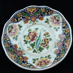 May include: A round ceramic bowl with a scalloped edge, featuring a white base decorated with hand-painted floral patterns in shades of yellow, red, green, and dark blue. The central design is a bouquet surrounded by a circular border.