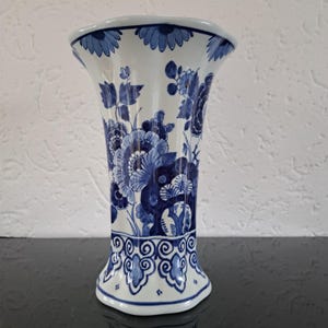 May include: A white ceramic vase with a flared top and a detailed blue floral pattern. The vase has a wide base and a scalloped rim. The blue floral design covers the entire surface of the vase, creating a classic, elegant look.