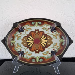 May include: An oval ceramic serving dish with a brown rim and a colorful floral design. The design features a central flower with yellow, red, and brown petals, surrounded by smaller flowers and leaves in shades of green, blue, and red.