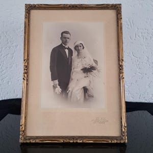 May include: A black and white photograph of a couple in a wedding portrait. The couple is dressed in formal attire, the woman is wearing a white dress and veil, and the man is wearing a tuxedo. The photograph is framed in a gold frame.