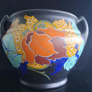 Może przedstawiać: Matowe czarne naczynie ceramiczne z dwoma bocznymi uchwytami, ozdobione żywym kwiatowym wzorem w odcieniach pomarańczu, żółci, błękitu i jasnej zieleni. Wzór zawiera stylizowane liście i płatki z wyraźnymi konturami na ciemnym tle.